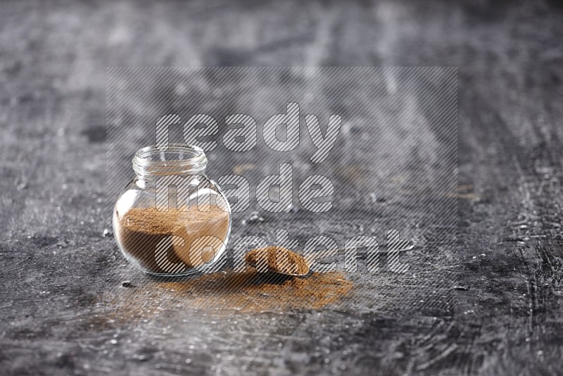 Herbal glass jar full of cinnamon powder and a metal spoon full of powder on textured black background