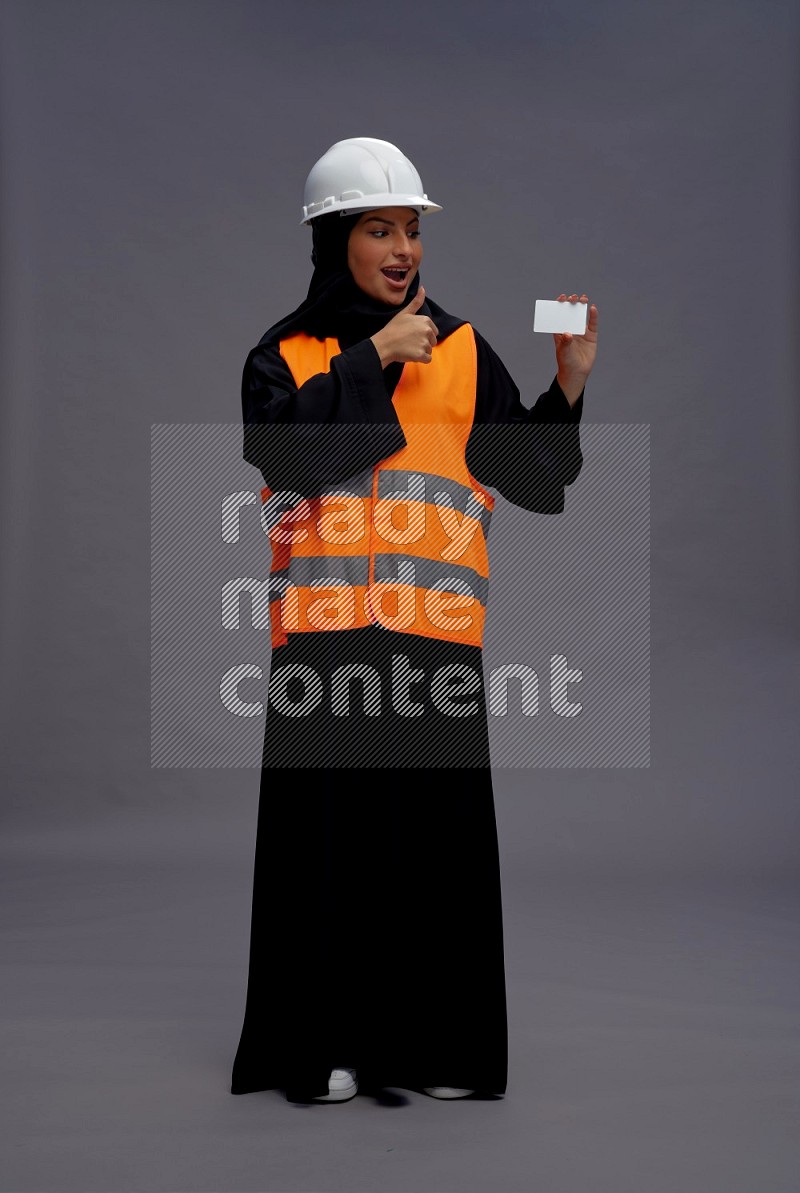 Saudi woman wearing Abaya with engineer vest standing holding ATM card on gray background