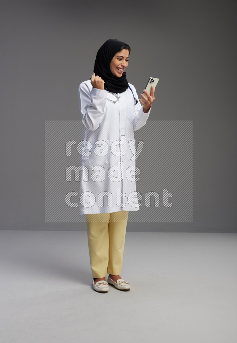 Saudi woman wearing lab coat with stethoscope standing texting on phone on Gray background