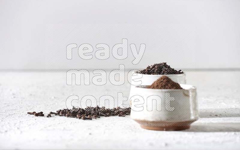 2 Beige ceramic bowls, one full of cloves and the other full of powder on textured white flooring