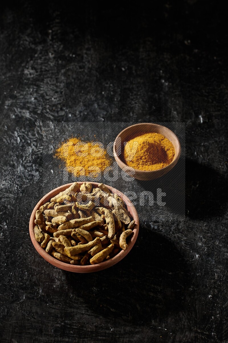 2 wooden bowls, one full of turmeric powder and the other full of dried turmeric whole fingers on a textured black flooring