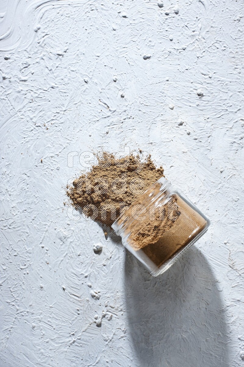 A flipped glass jar full of allspice powder and powder spilled out of it on a textured white flooring