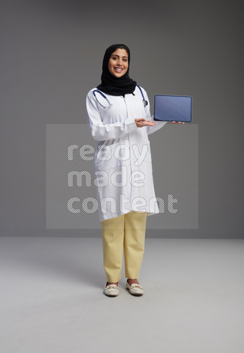Saudi woman wearing lab coat with stethoscope standing showing tablet to camera with sign in the back on Gray background