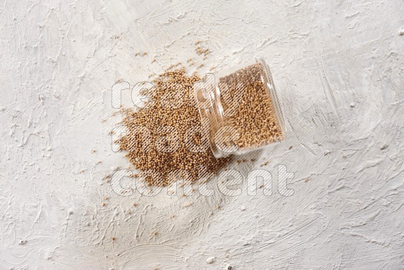 A glass jar full of mustard seeds and jar is flipped with seeds spread out on a textured white flooring