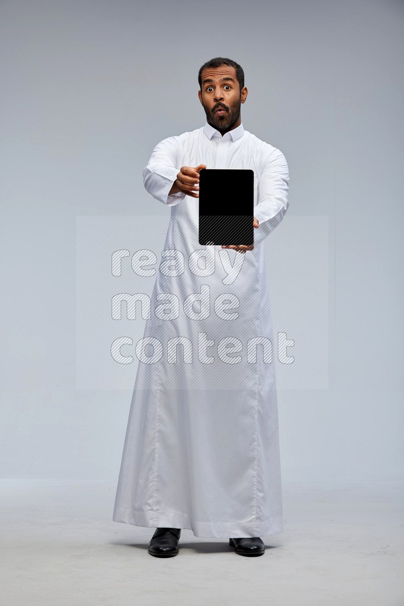 Saudi man wearing Thob standing showing tablet to camera on Gray background