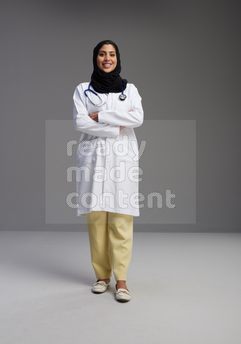 Saudi woman wearing lab coat with stethoscope standing with crossed arms on Gray background
