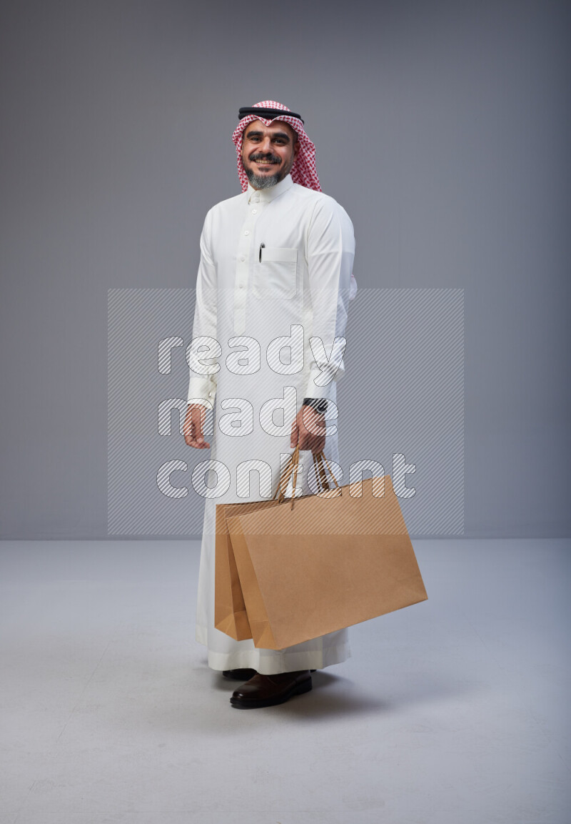 Saudi man Wearing Thob and red Shomag standing holding shopping bag on Gray background