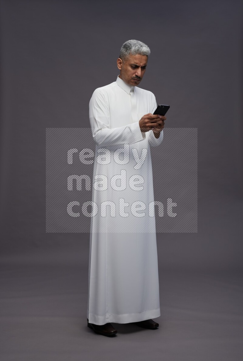 Saudi man wearing thob standing texting on phone on gray background