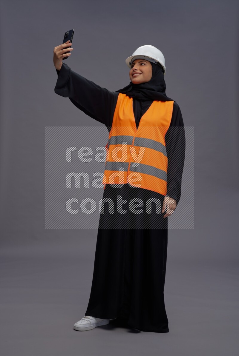 Saudi woman wearing Abaya with engineer vest standing taking selfie on gray background