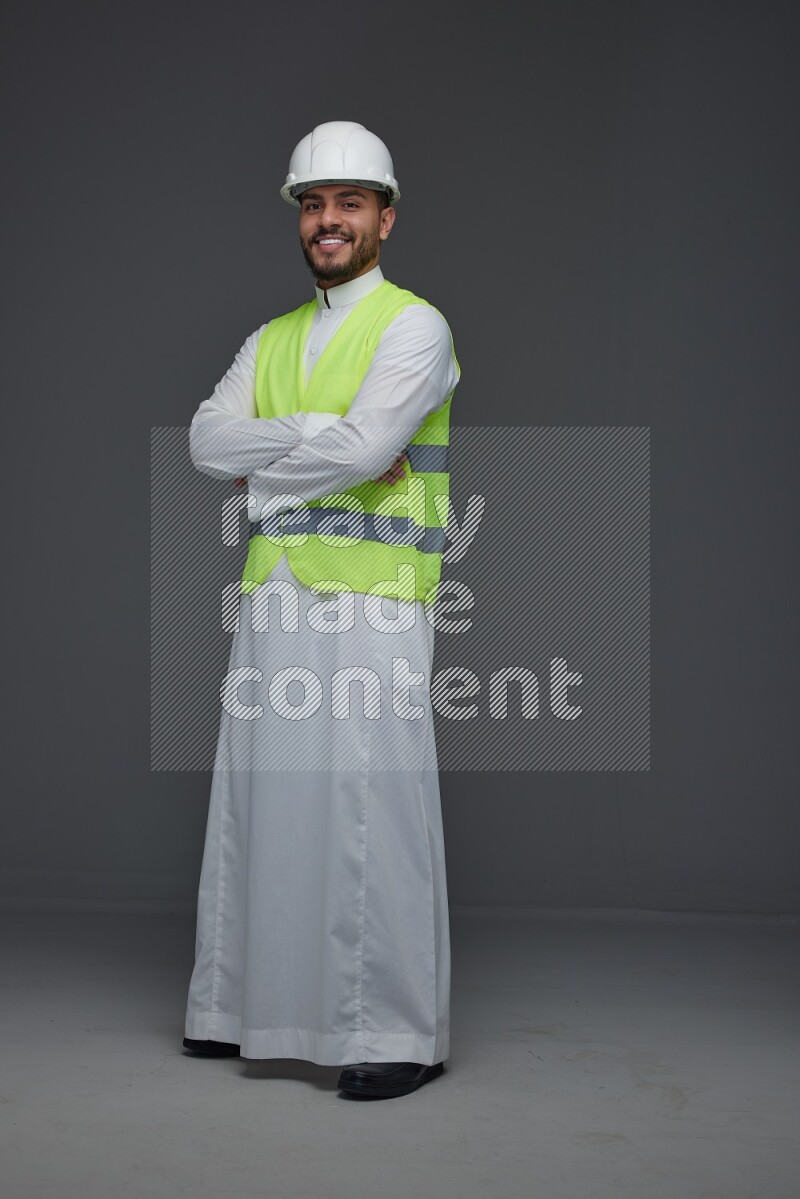 A Saudi man wearing Thobe with a yellow safety vest and white helmet standing and crossing his hands eye level on a gray background