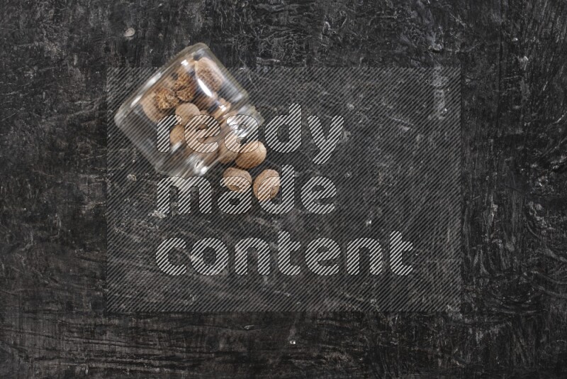 A glass jar full of nutmeg flipped and the seeds spread out on a textured black flooring