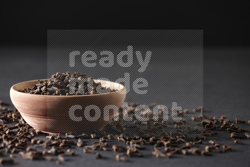 A wooden bowl full of cloves with spreaded whole cloves on a black flooring