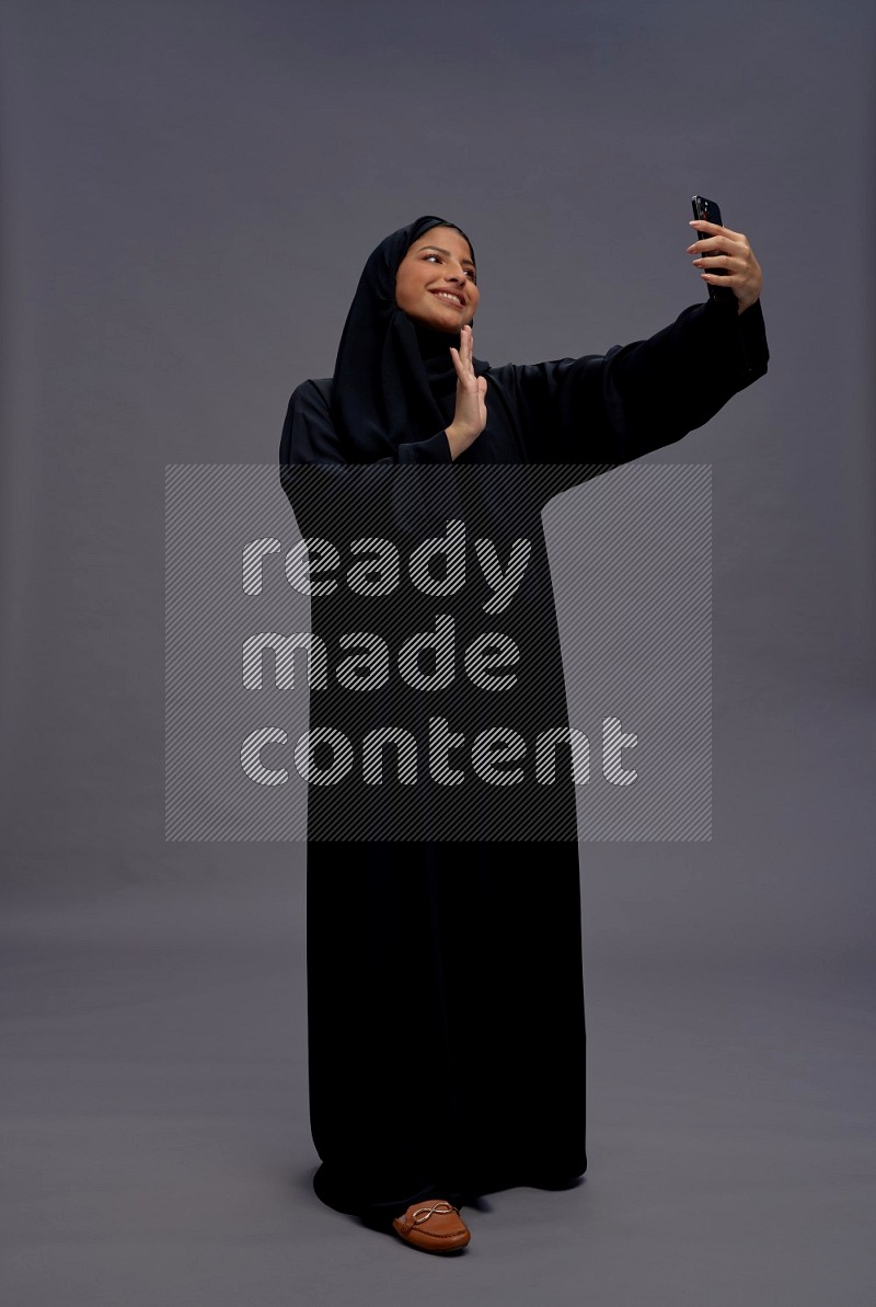 Saudi woman wearing Abaya standing taking selfie on gray background