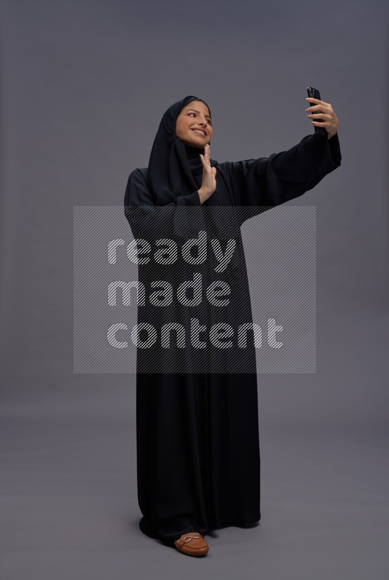 Saudi woman wearing Abaya standing taking selfie on gray background