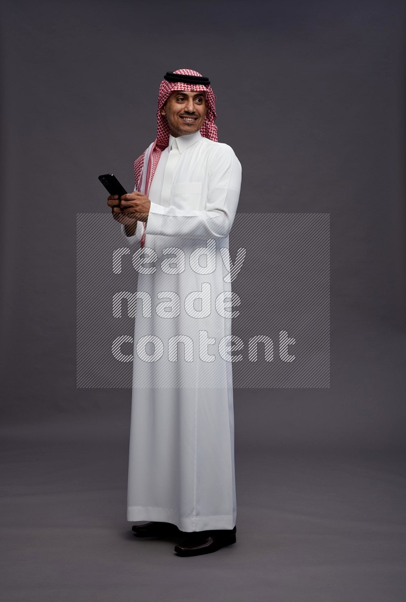 Saudi man wearing thob and shomag standing texting on phone on gray background