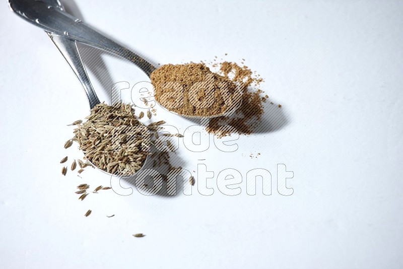 2 metal spoons full of cumin seeds and powder on white flooring