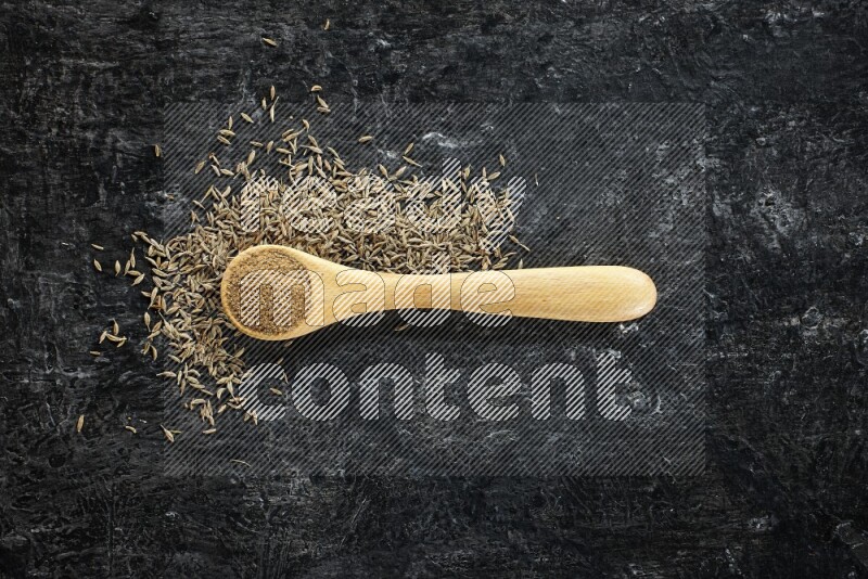 A wooden spoon full of cumin powder and cumin seeds spreaded on textured black flooring