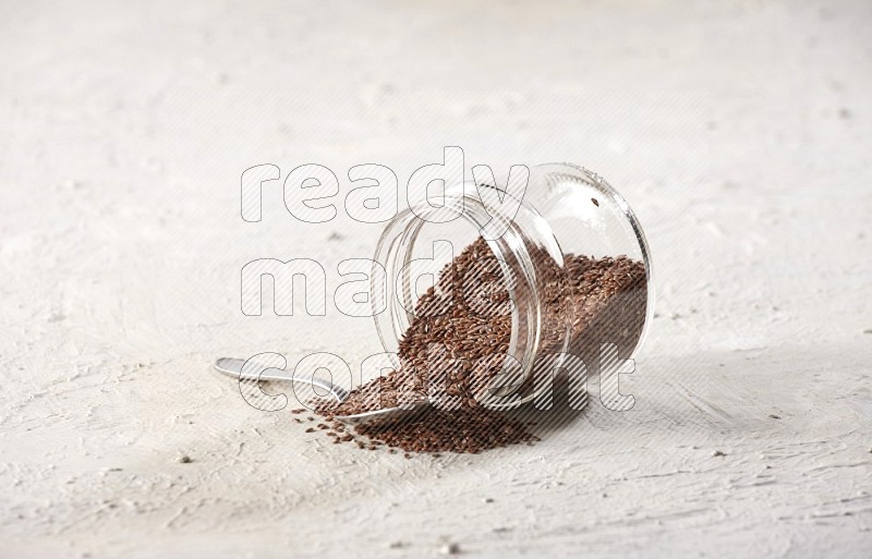 A glass jar full of flax flipped and flax spreaded out with metal spoon full of the seeds on a textured white flooring in different angles