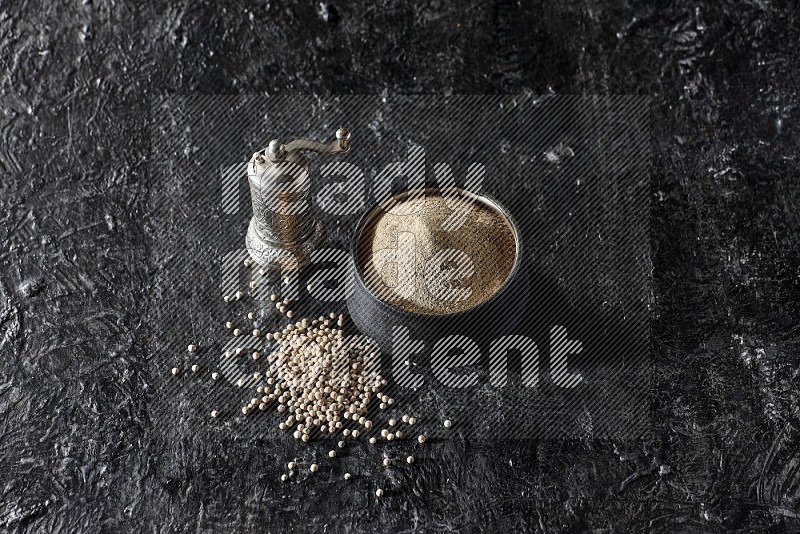 Black pottery bowl full of white pepper powder with pepper beads and metal pepper grinder on textured black flooring