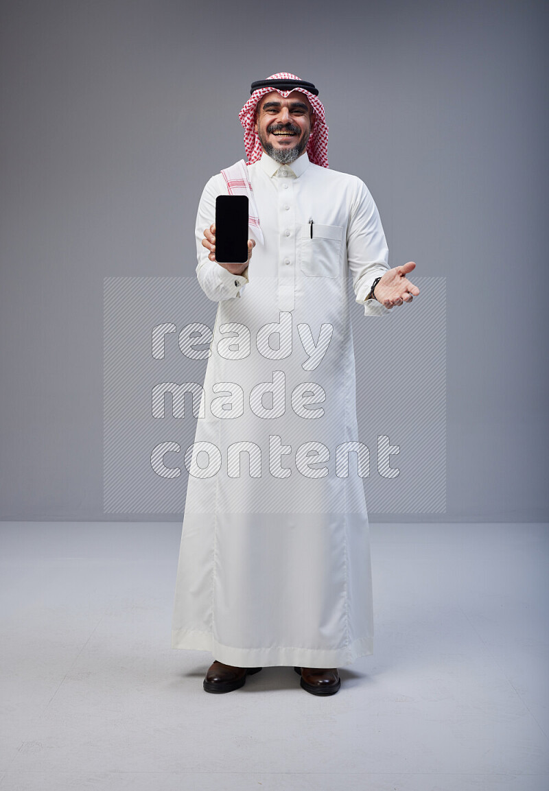 Saudi man Wearing Thob and red Shomag standing showing phone to camera on Gray background