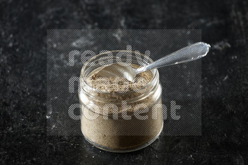 A glass jar and metal spoon full of cardamom powder on textured black flooring