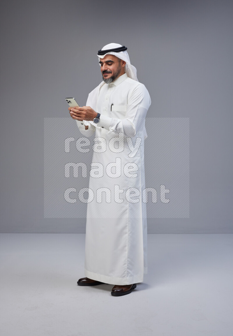 Saudi man Wearing Thob and white Shomag standing texting on phone on Gray background