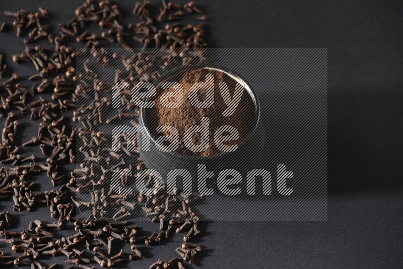 A black pottery bowl full of cloves powder and cloves whole grains on a black flooring