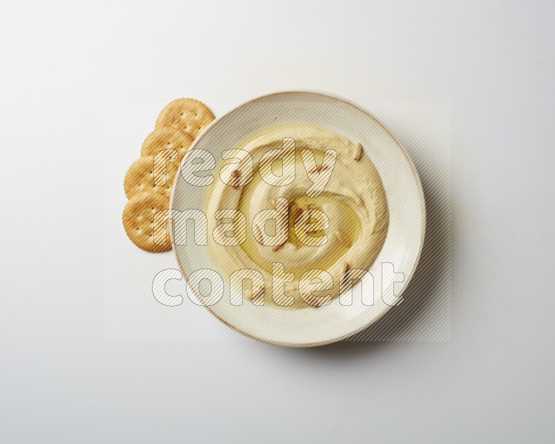 Hummus in a pottry plate garnished with pine nuts on a white background