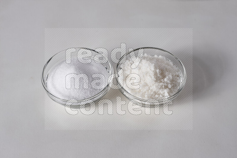 2 glass bowls one is filled with table salt and the other with coarse sea salt on white background