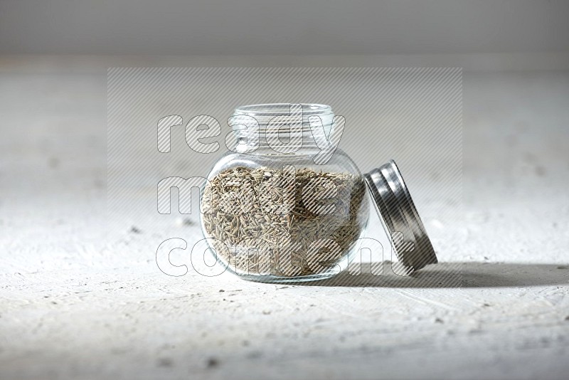 A glass spice jar full of cumin seeds on textured white flooring