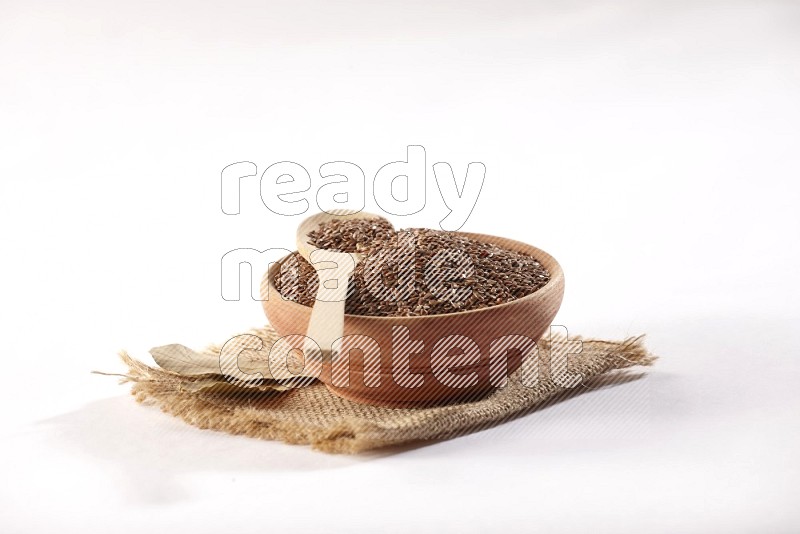 A wooden bowl full of flax seeds and a wooden spoon full of the seeds on burlap fabric on a white flooring