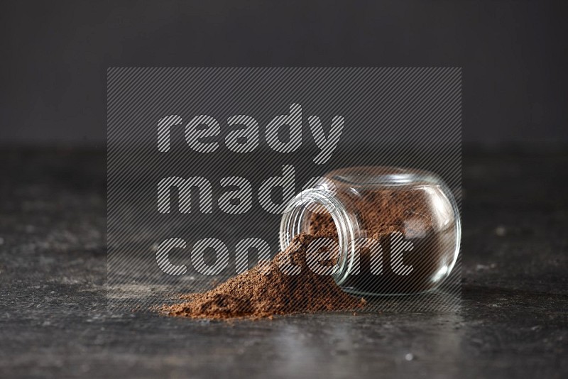 A flipped glass spice jar full of cloves powder and powder came out of it on textured black flooring