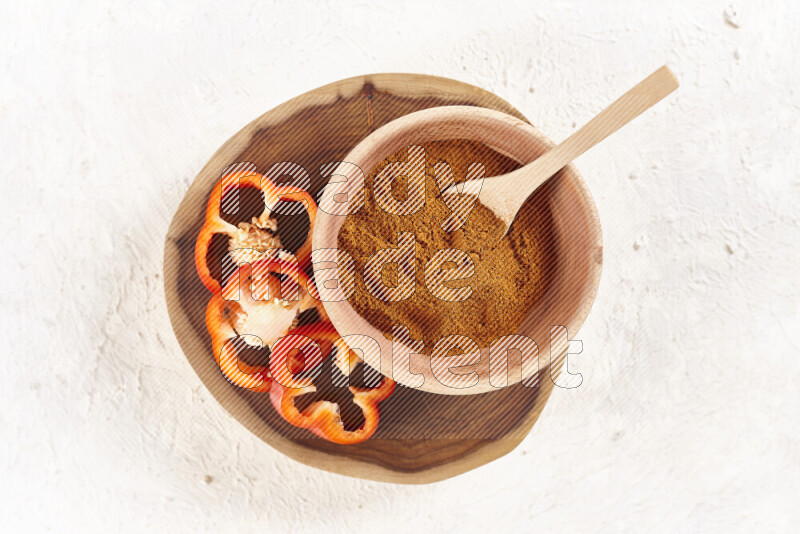 A wooden bowl full of ground paprika powder and sliced red bell pepper beside it all on a wooden tray on white background