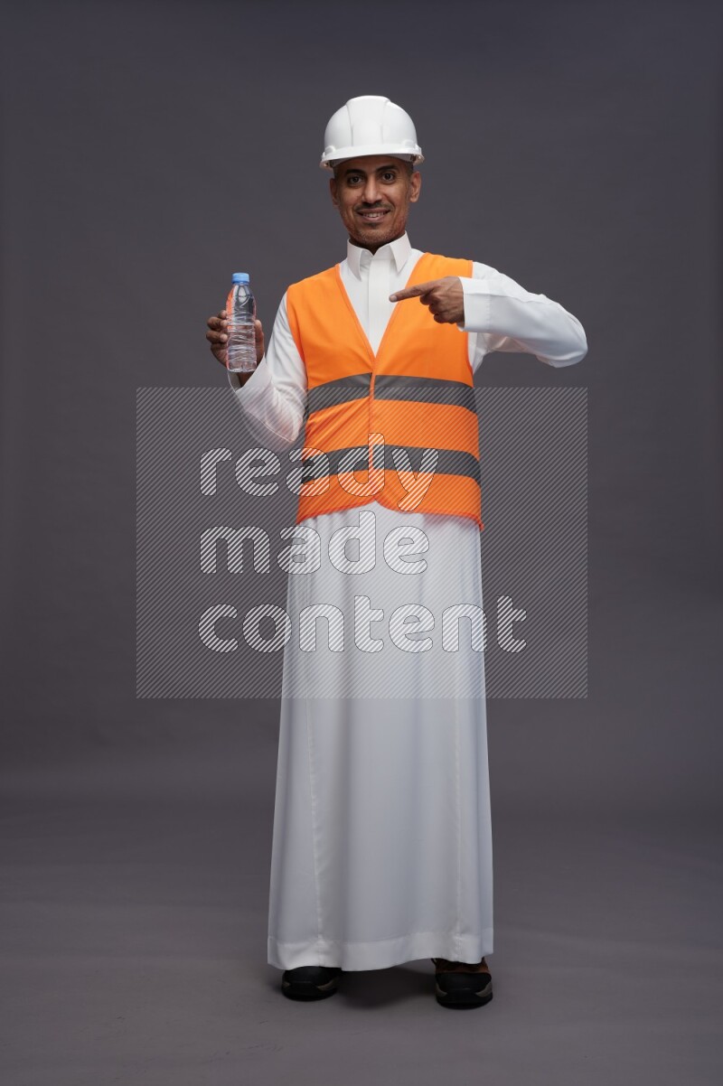 Saudi man wearing thob with engineer vest standing drinking water on gray background