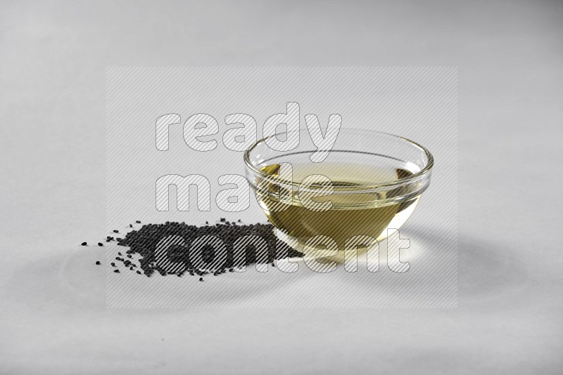 A glass bowl full of black seeds oil with black seeds beside it on a white flooring