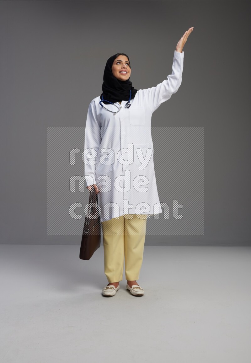 Saudi woman wearing lab coat with stethoscope standing holding bag on Gray background