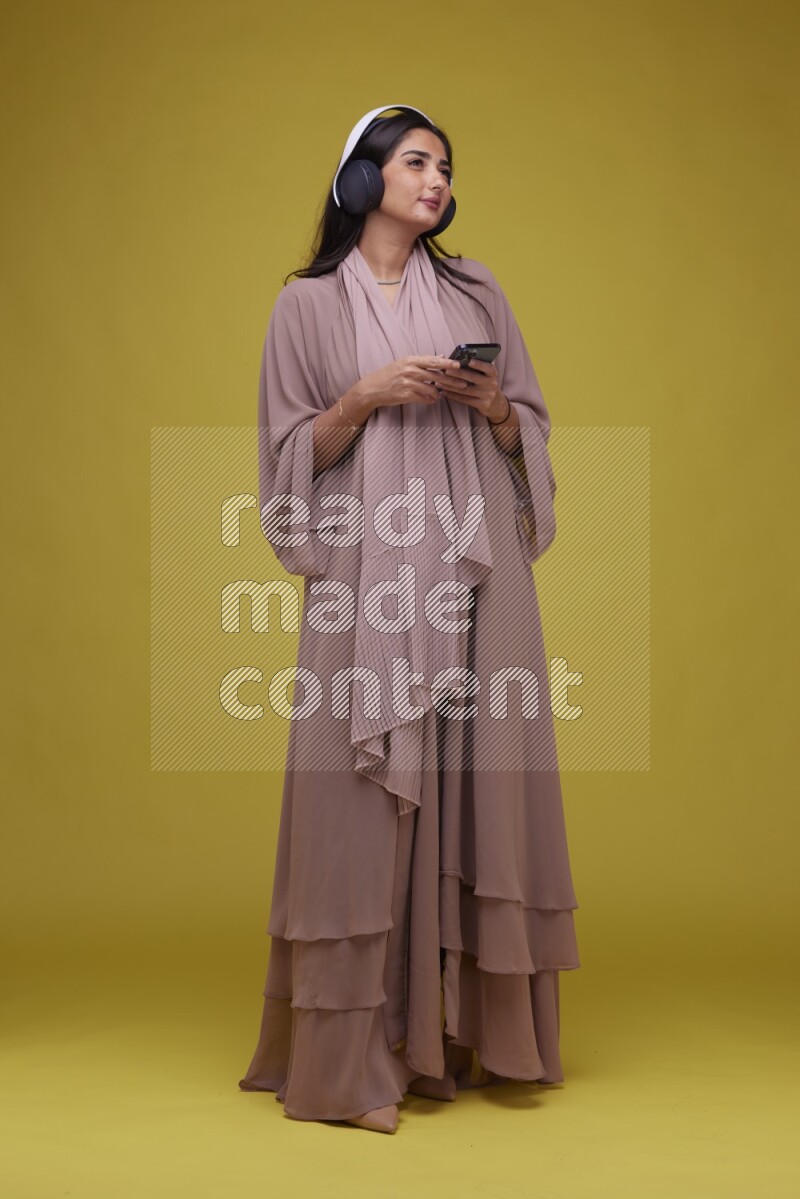 A Saudi woman Listing to Music on a Yellow Background wearing Brown Abaya