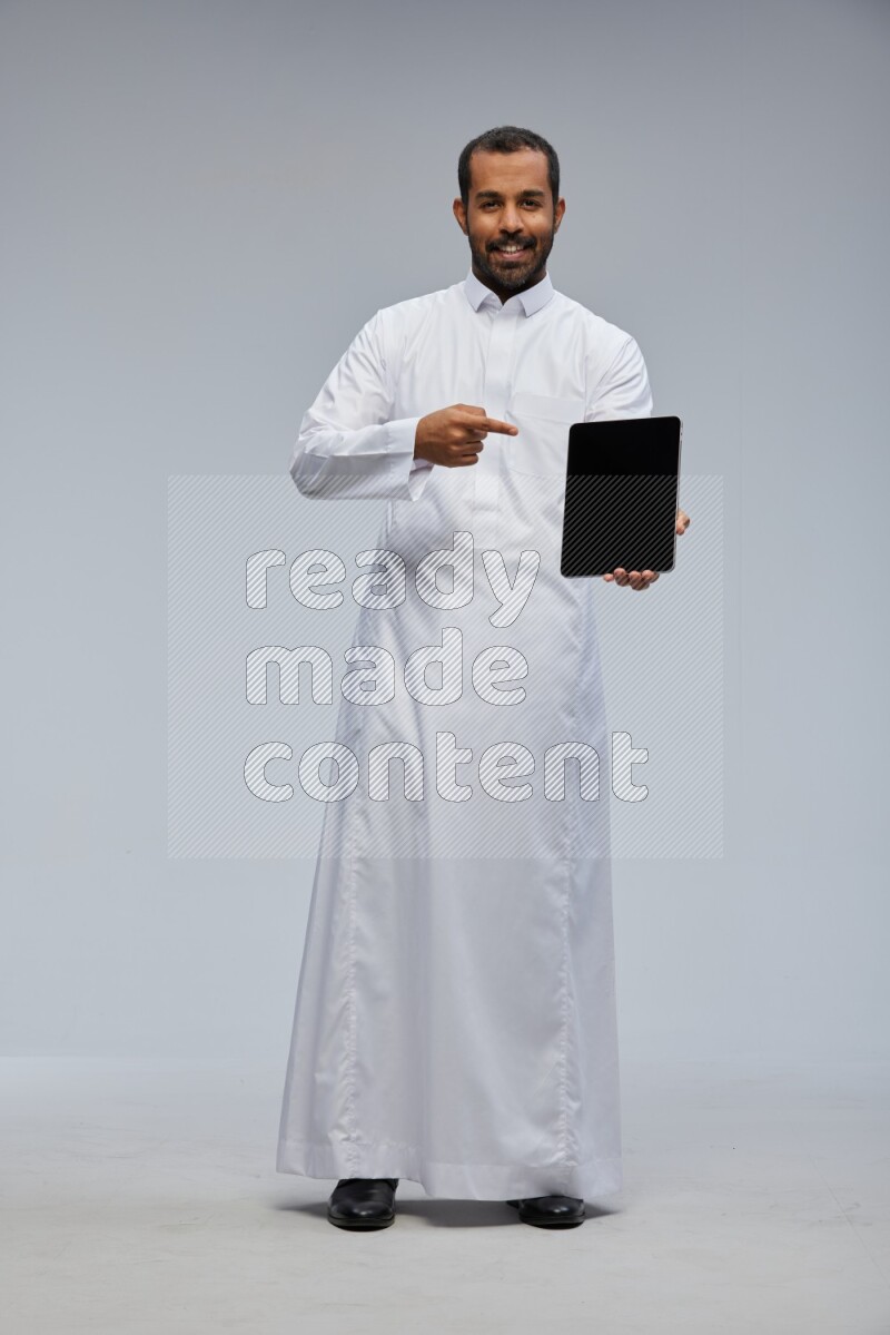 Saudi man wearing Thob standing showing tablet to camera on Gray background