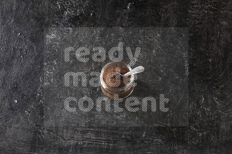 A glass jar full of cloves powder with a metal spoon on a textured black flooring