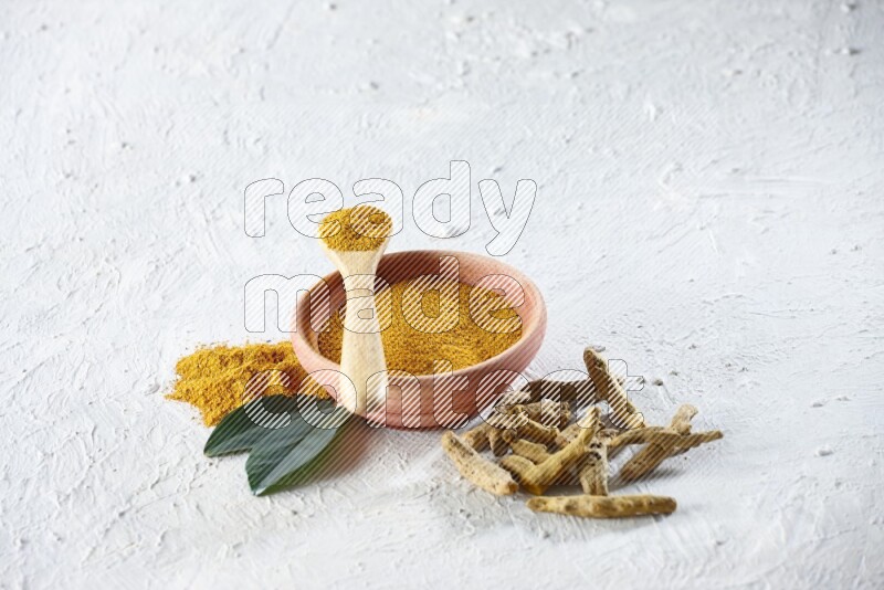 A wooden bowl and wooden spoon full of turmeric powder with dried turmeric fingers beside it on textured white flooring