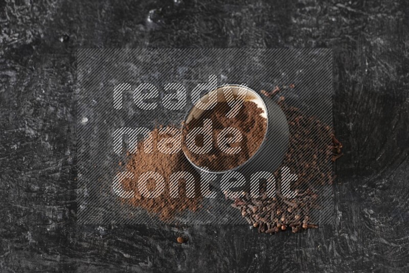 A black pottery bowl full of cloves powder spilled out of it with whole grains on a textured black flooring