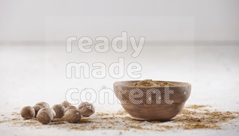 A wooden bowl full of nutmeg powder with whole seeds and sprinkled powder beside it on a textured white flooring