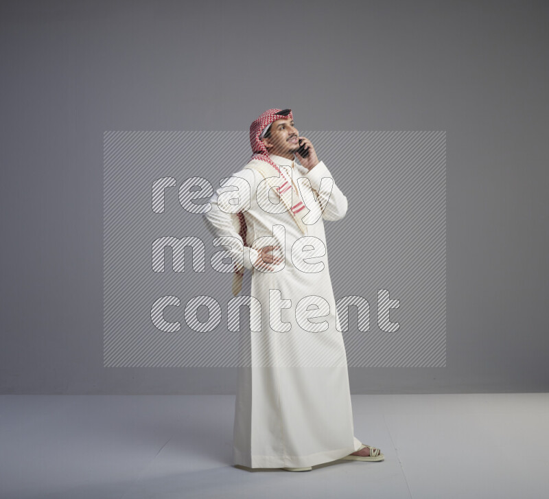 A Saudi man standing wearing thob and red shomag talking on phone on gray background
