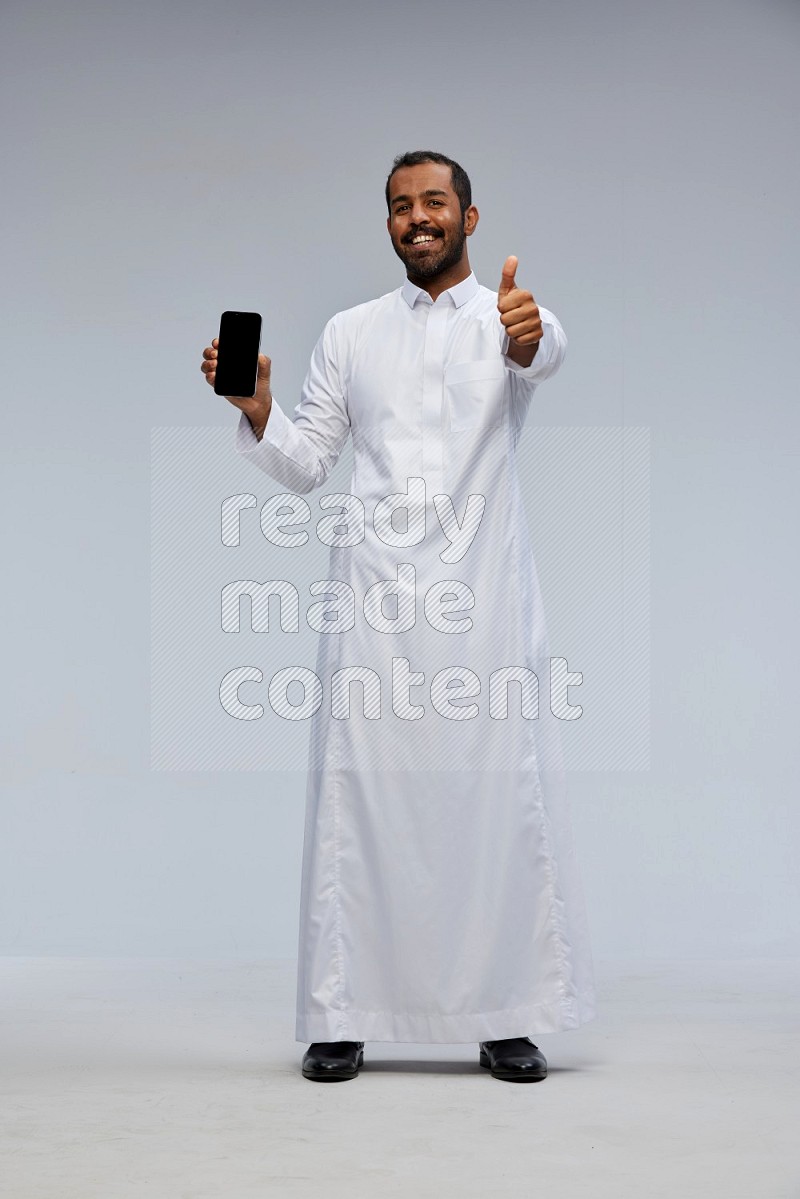 Saudi man wearing Thob standing showing phone to camera on Gray background