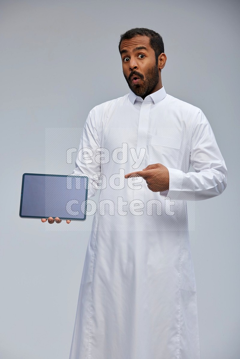 Saudi man wearing Thob standing showing tablet to camera on Gray background