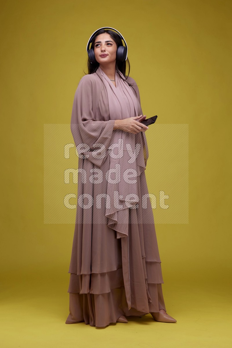 A Saudi woman Listing to Music on a Yellow Background wearing Brown Abaya