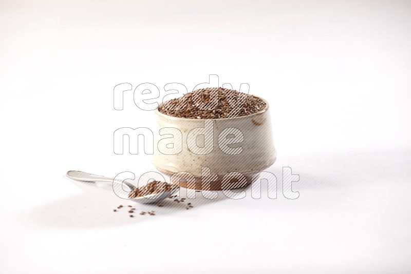 A pottery beige bowl and a metal spoon full of flax seeds on a white flooring