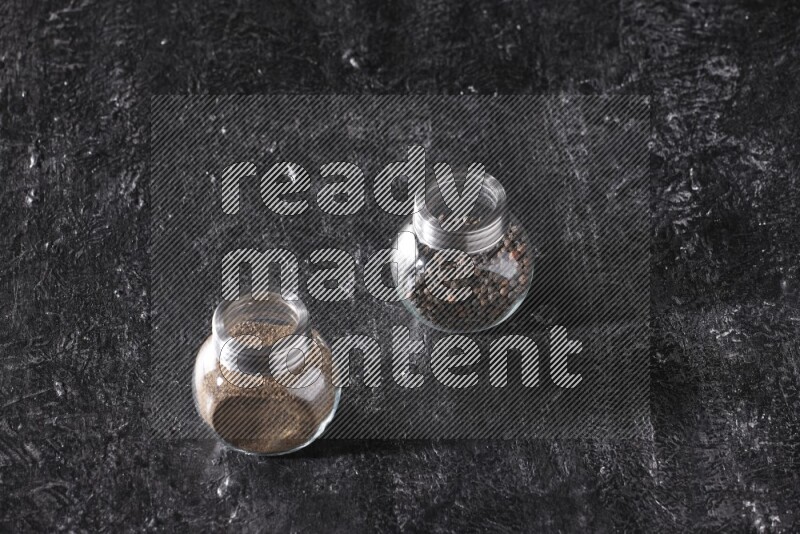 2 glass spice jars full of black pepper powder and black pepper beads on textured black flooring