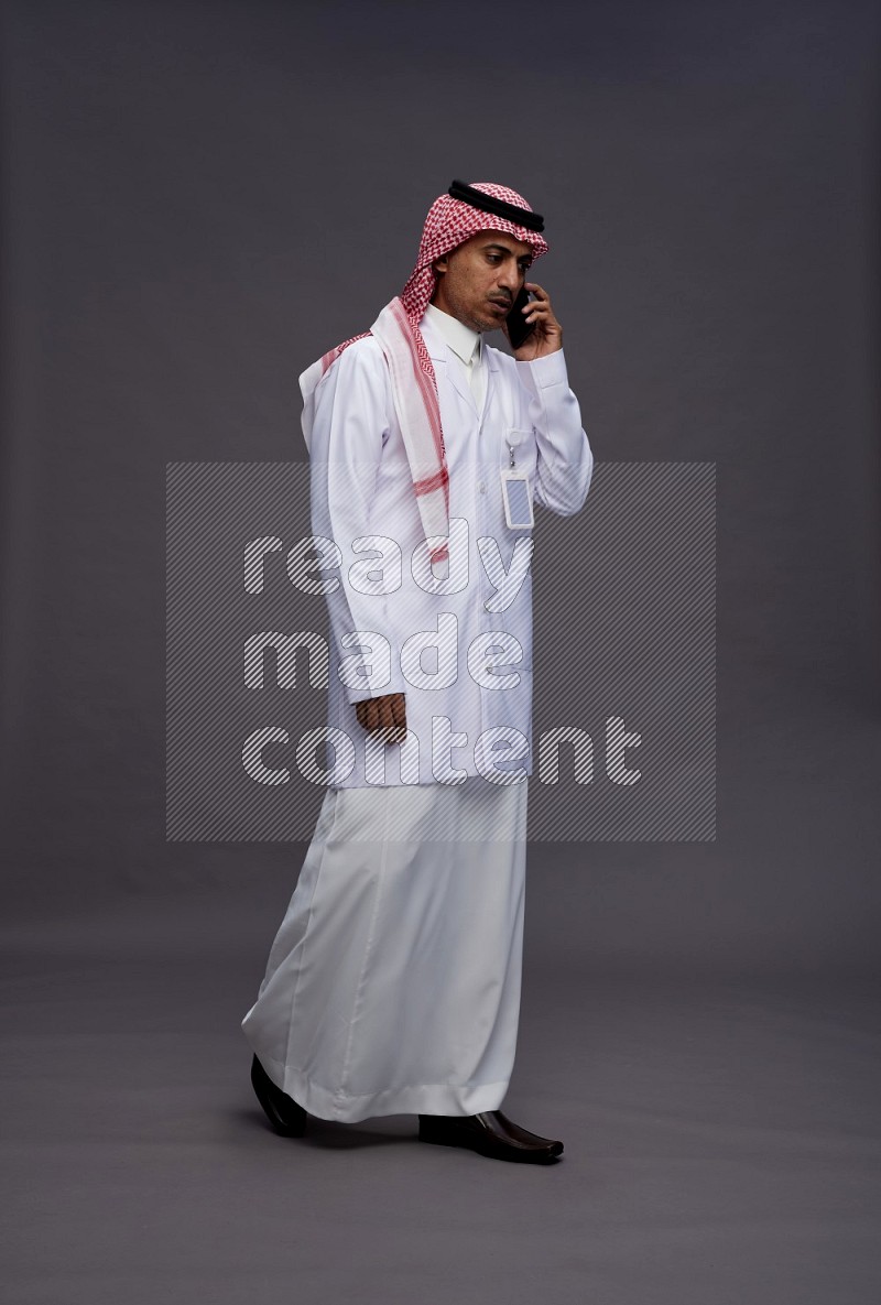 Saudi man wearing thob with lab coat and shomag with pocket employee badge standing talking on phone on gray background