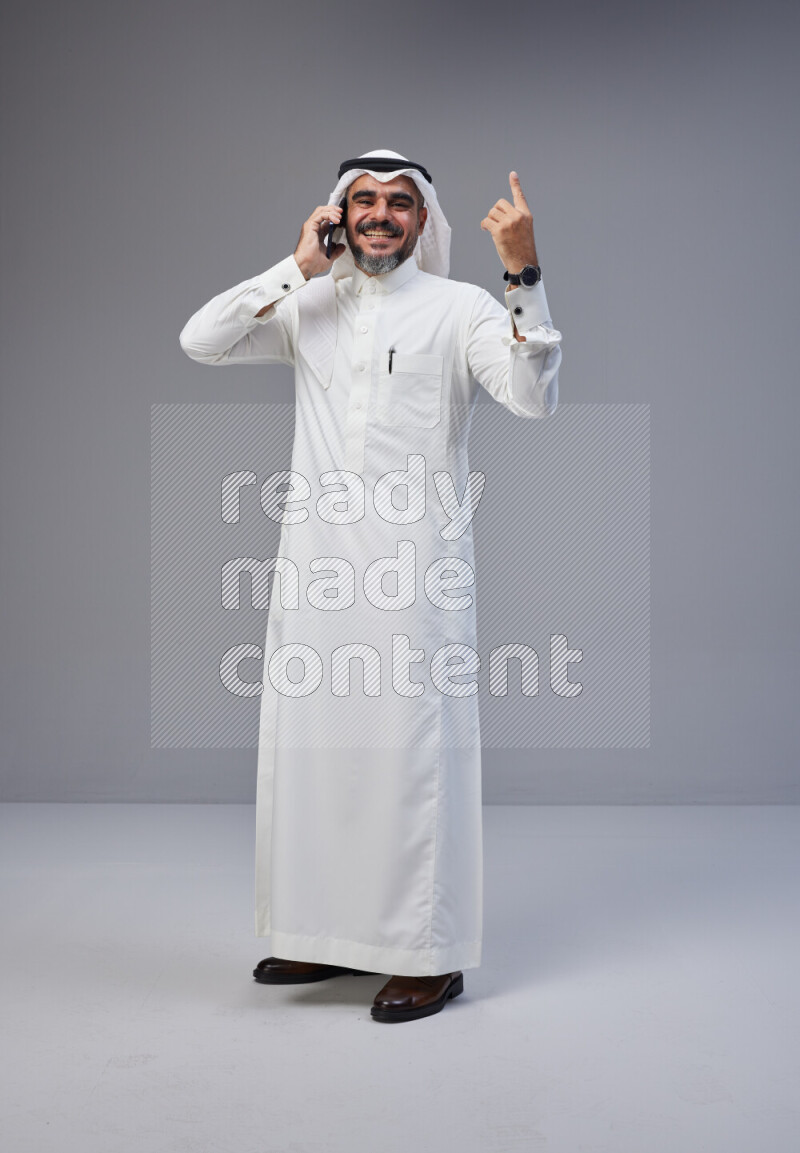 Saudi man Wearing Thob and white Shomag standing talking on phone on Gray background
